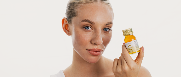 Kobieta w blond włosach o promiennej cerze trzyma butelkę kolagenu do picia Primabiotic.