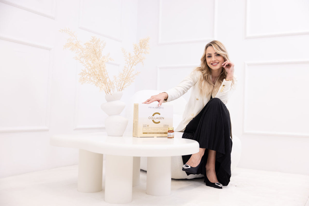 Dr Wanda Baltaza uśmiechnięta, prezentuje Primabiotic Collagen do picia – eleganckie opakowanie z buteleczką suplementu na stole