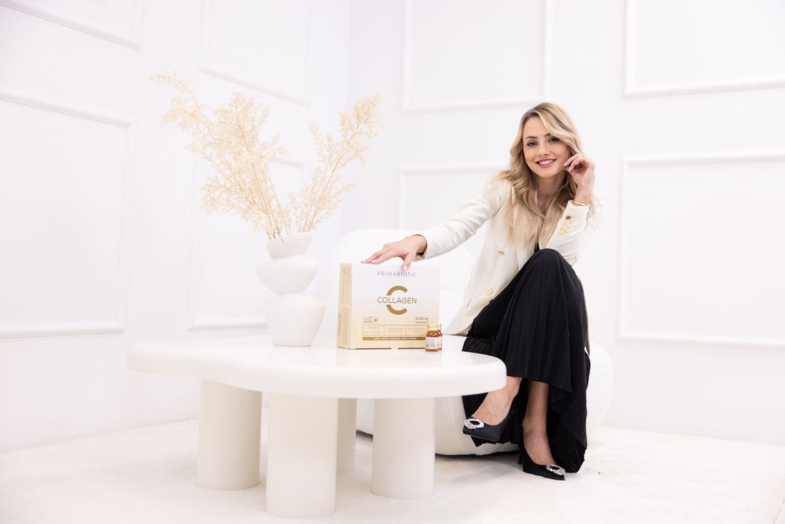 Dr Wanda Baltaza uśmiechnięta, prezentuje Primabiotic Collagen do picia – eleganckie opakowanie z buteleczką suplementu na stole