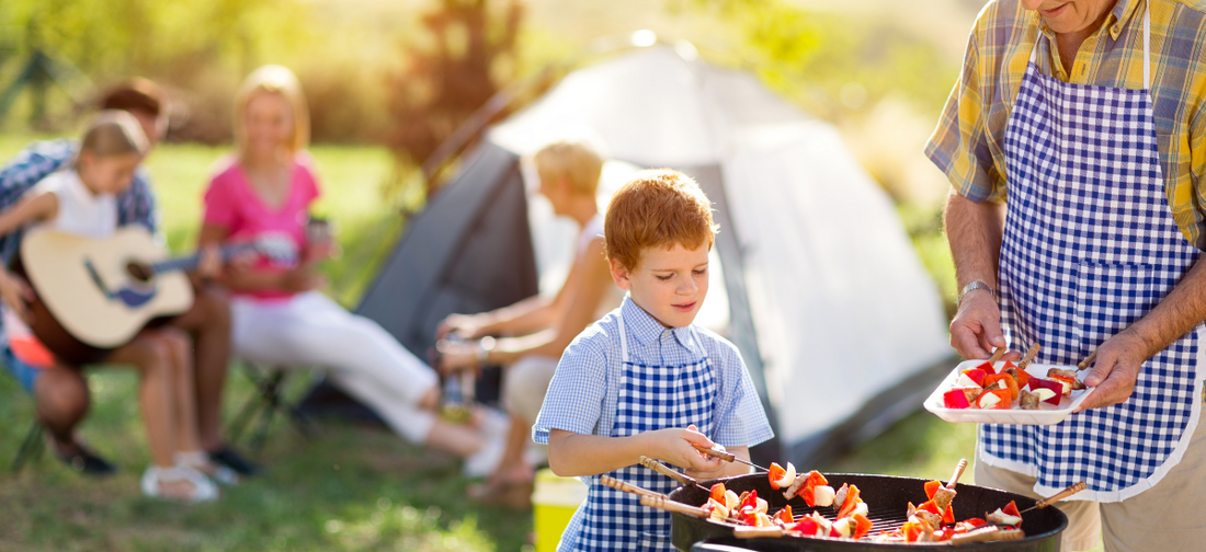 Chłopiec w fartuszku przyrządza szaszłyki na grillu obok dorosłego, w tle namiot i rodzina odpoczywająca na biwaku