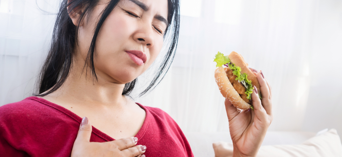 Kobieta trzyma burgera i dotyka klatki piersiowej z grymasem bólu, objawy refluksu po ciężkostrawnym posiłku