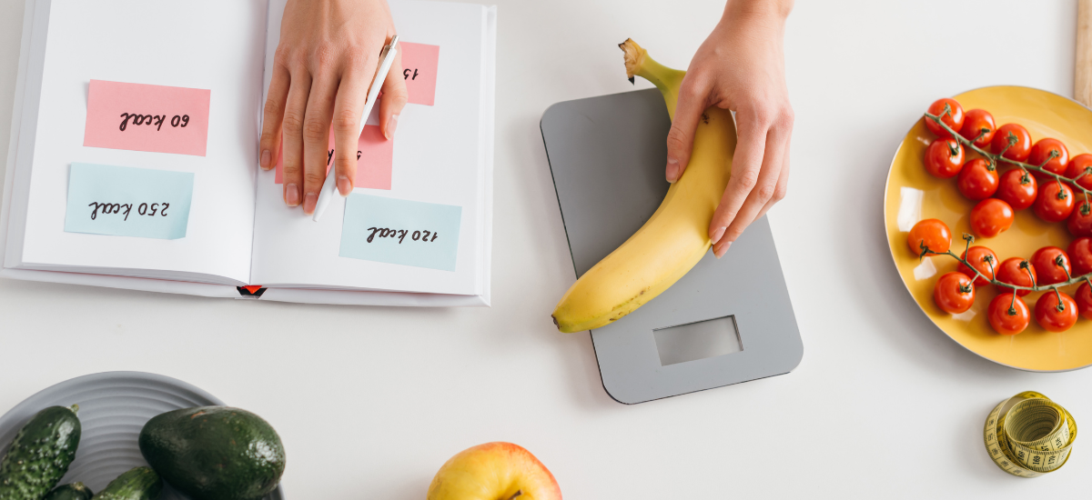Kobieta waży banana leżącego obok warzyw obliczając w zeszycie deficyt kaloryczny