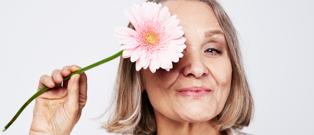 Uśmiechnięta kobieta w okresie menopauzy trzymająca różowy kwiat nad okiem, prezentująca swoją promienną skórę i piękne włosy po suplementacji kolagenu do picia Primabiotic