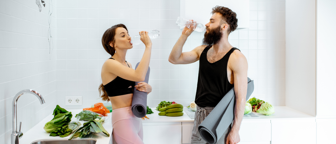 Kobieta i mężczyzna w sportowych strojach piją wodę z butelek. Oboje trzymają maty do ćwiczeń. Na stoliku w tle widać wiele warzyw.